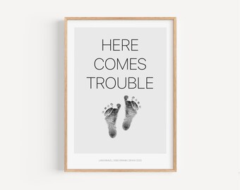 May include: A framed print with a gray background and black text that reads "Here Comes Trouble".  Two black and white baby footprints are below the text.  The text "Liam Samuel | 3000 grams | 28 Nov 2020" is at the bottom of the print.