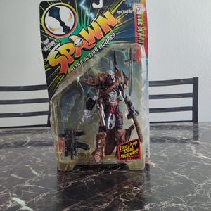 May include: A Spawn action figure with four drop dead weapons. The figure is in a plastic package with a red, white, and black color scheme. The figure is wearing a black and red outfit with a skull on the chest. The figure is holding a sword and a gun. The package has the words "Spawn Ultra Action Figures" on it.