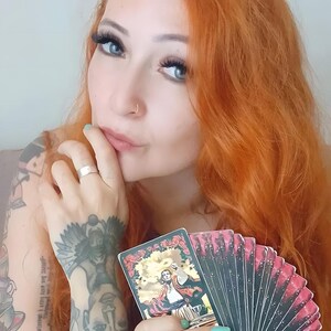 May include: A person with red hair and blue eyes holds a deck of tarot cards. The cards have a red and black design with a central illustration. The person has tattoos on their arm and hand, and is wearing a silver ring.
