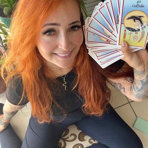 May include: A person holding a deck of tarot cards, with the "Golfinho" card visible. The cards have a blue and white design. The person is wearing a black shirt and pants. The person has red hair and is smiling.