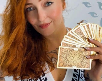 May include: A woman with long, red hair holds a fan of gold tarot cards with intricate designs. She is wearing a white top with a black and white geometric pattern.