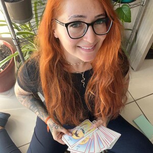 May include: A person holding a deck of tarot cards. The cards have colorful illustrations with a crescent moon and stars. The person is wearing black glasses, a black shirt, and a silver necklace. The person has long red hair.