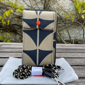 Könnte beinhalten: Eine beige und marineblau gemusterte Geldbörse mit passendem Riemen. Die Geldbörse hat eine Klappe mit orangefarbenem Knopfverschluss und einen Reißverschluss. Der Riemen ist schwarz und weiß, und die Geldbörse liegt auf einem weißen Tuch.