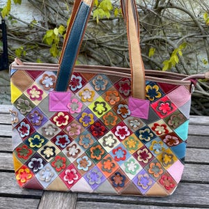 Pode incluir: Uma sacola tote colorida em patchwork com um design floral. A sacola apresenta uma variedade de cores, incluindo vermelho, rosa, amarelo e azul, com um padrão geométrico. A sacola tem duas alças e um fecho de zíper.