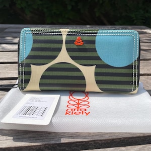 May include: A rectangular zip-around wallet featuring a dark green and black horizontal striped pattern with large cream and light blue circular accents. An orange flower-shaped emblem is centered on the front. A white dust bag with the text Orla Kiely is included.