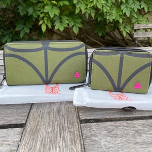 May include: Two olive green wallets with a gray leaf design and a pink floral emblem. The wallets are zippered and rest on white fabric with a logo. The background features green foliage and a wooden surface.