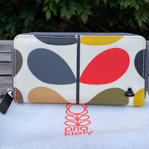 May include: A rectangular wallet with a colorful leaf pattern in red, yellow, blue, and brown on a white background. The wallet has a gray zipper and a black tag. The brand name "orla kiely" is visible.