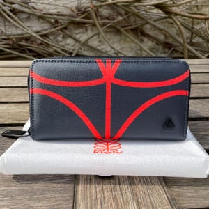 May include: A dark gray wallet with a red graphic design. The wallet has a zipper closure and a small logo on the front. The wallet is sitting on a white cloth.