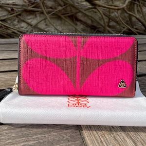 May include: A pink wallet with a brown floral pattern. The wallet has a black zipper and a gold-colored zipper pull. The wallet is on a white cloth with the brand name "Orla Kiely" printed on it.