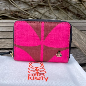 May include: A pink and brown patterned zip-around wallet with a gold zipper and a small gold emblem. The wallet is on top of a white cloth bag with the brand name "orla kiely" printed on it.