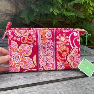 Puede incluir: Bolsa de cosméticos acolchada y rectangular con un vibrante estampado floral en tonos rosa, naranja y amarillo. La bolsa tiene cierre de cremallera y un pequeño lazo. Una etiqueta verde cuelga. Aproximadamente 20 cm de ancho.