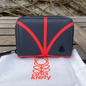 May include: A small, rectangular wallet in a dark gray fabric with a red geometric design. The wallet has a black zipper and a small, black logo. A white bag with the brand name "orla kiely" in orange is in front of the wallet.