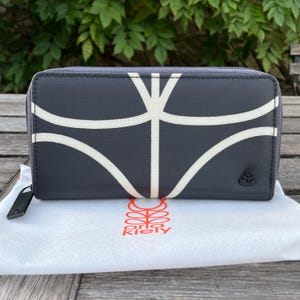 May include: A dark gray wallet with a white abstract design. The wallet has a zipper closure and a small logo on the front. The wallet is on top of a white cloth with the brand name "Orla Kiely" in orange.