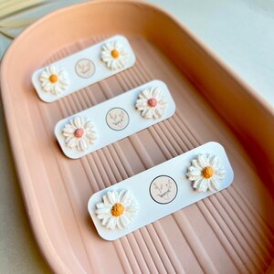 May include: Three pairs of white daisy flower stud earrings displayed on a pink tray. Each pair is on a white card with a small floral logo.