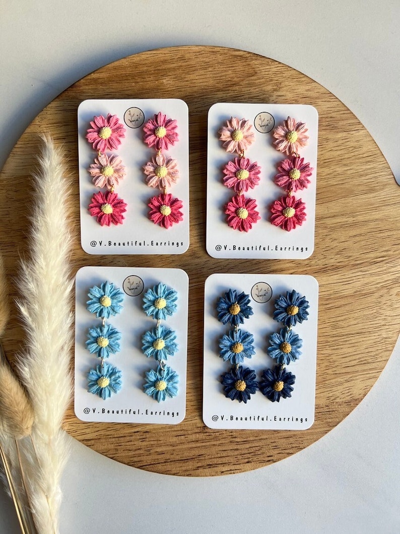 May include: Four pairs of dangle earrings featuring three layered flower charms. The earrings are in shades of pink, blue, and navy blue. The flowers have yellow centers. The earrings are displayed on white cards with the text "@V.Beautiful.Earrings".