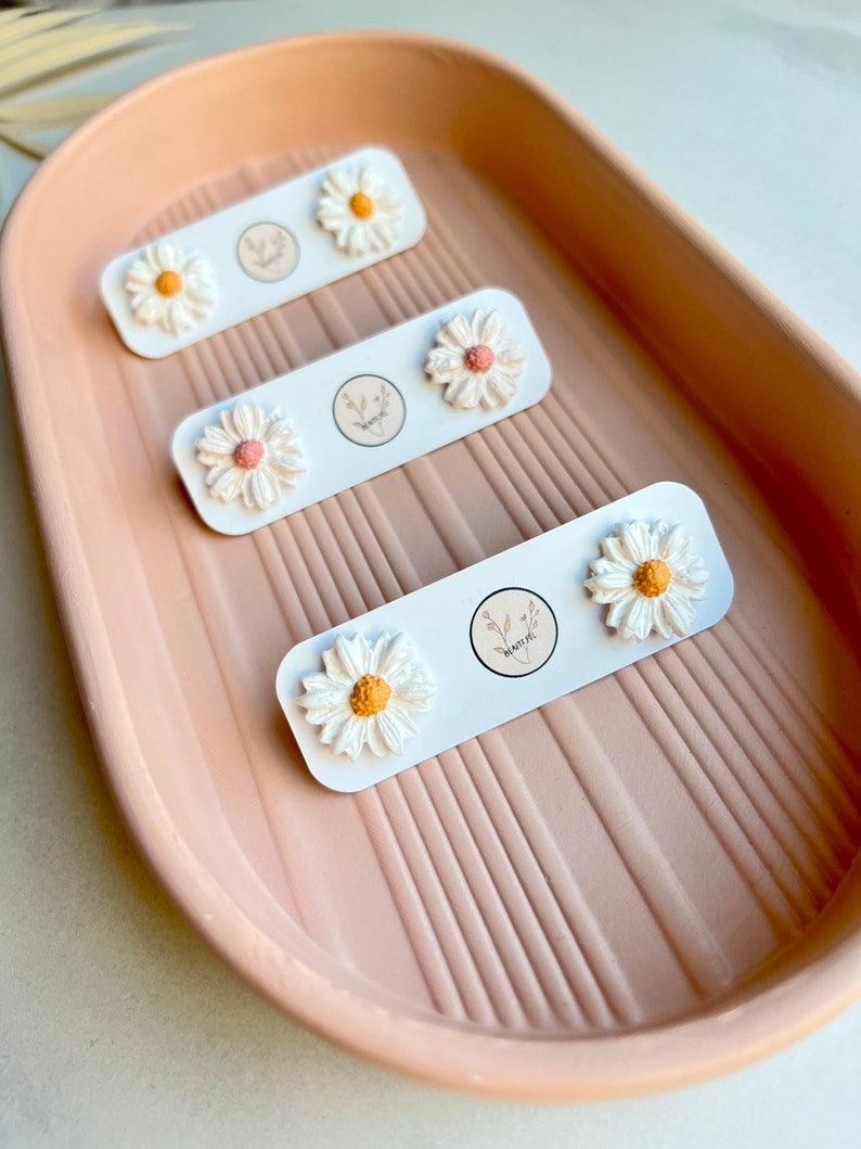 May include: Three pairs of white daisy flower stud earrings displayed on a pink tray. Each pair is on a white card with a small floral logo.