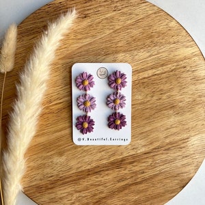 May include: A pair of dangle earrings with three purple and yellow flowers on each earring. The earrings are on a white card with the text "@V.Beautiful.Earrings".