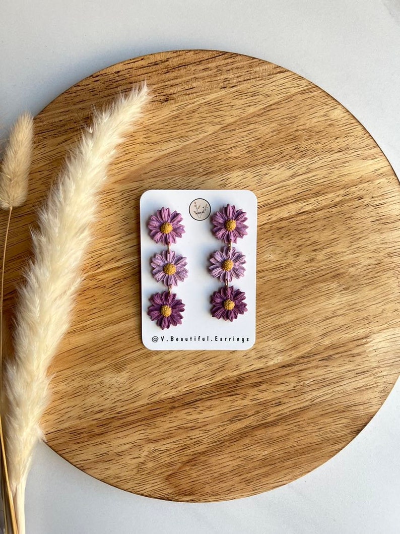 May include: A pair of dangle earrings with three purple and yellow flowers on each earring. The earrings are on a white card with the text "@V.Beautiful.Earrings".