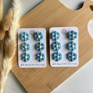May include: Two pairs of blue and yellow daisy earrings on white cards with the text "@V.Beautiful.Earrings" on each card.