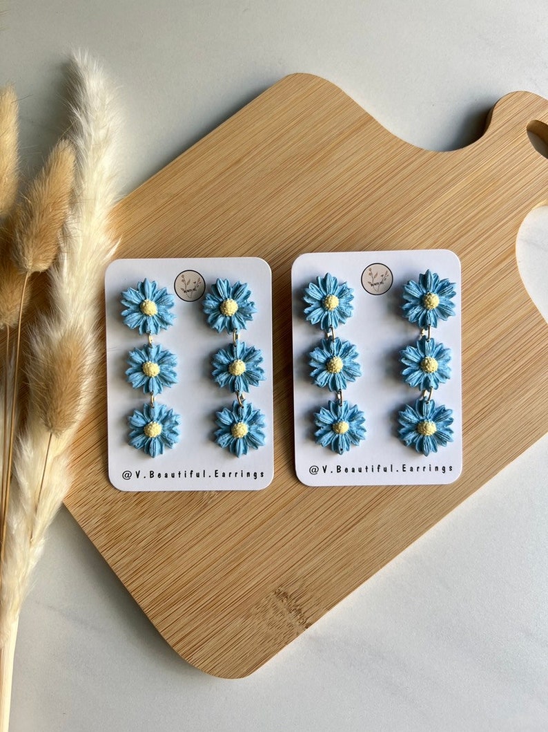 May include: Two pairs of blue and yellow daisy earrings on white cards with the text "@V.Beautiful.Earrings" on each card.