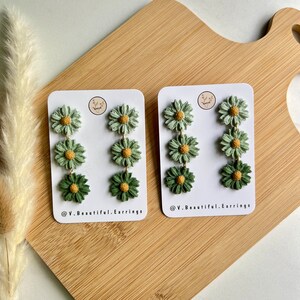 May include: A pair of green and yellow daisy flower dangle earrings. The earrings are on a white card with the text "@V.Beautiful.Earrings".