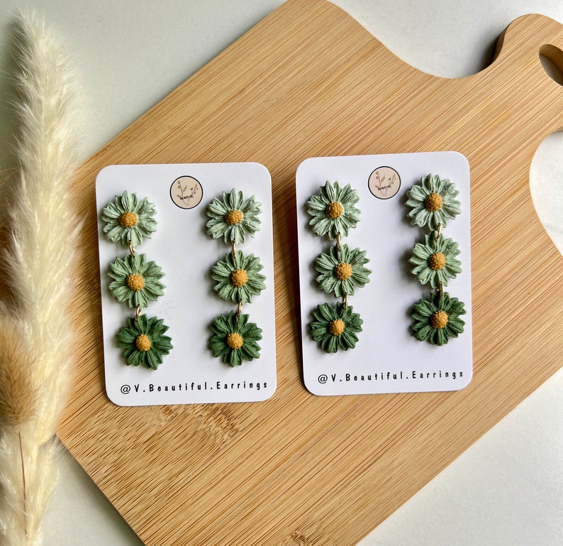 May include: A pair of green and yellow daisy flower dangle earrings. The earrings are on a white card with the text "@V.Beautiful.Earrings".