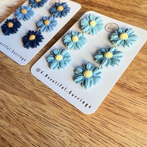 May include: A pair of blue and white flower dangle earrings. The earrings are made of polymer clay and have a light blue, medium blue, and dark blue flower design. The earrings are on a white card with the text "@V.Beautiful.Earrings".
