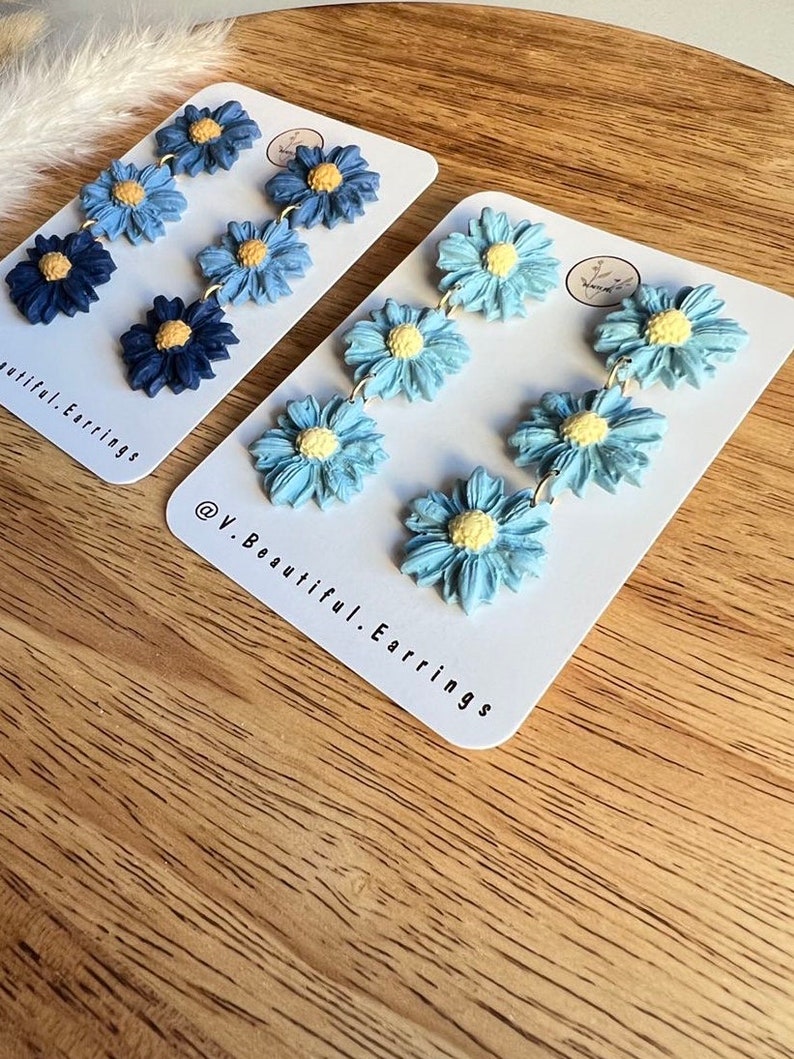 May include: A pair of blue and white flower dangle earrings. The earrings are made of polymer clay and have a light blue, medium blue, and dark blue flower design. The earrings are on a white card with the text "@V.Beautiful.Earrings".