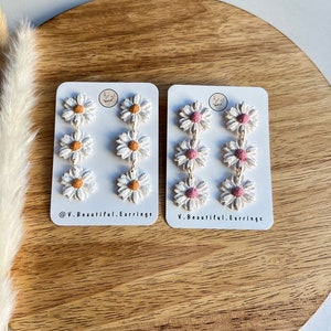 May include: Two pairs of daisy earrings on white cards. The earrings are made of white polymer clay with a pink or orange center. The cards have the text "@V.Beautiful.Earrings" and "V.Beautiful.Earrings".