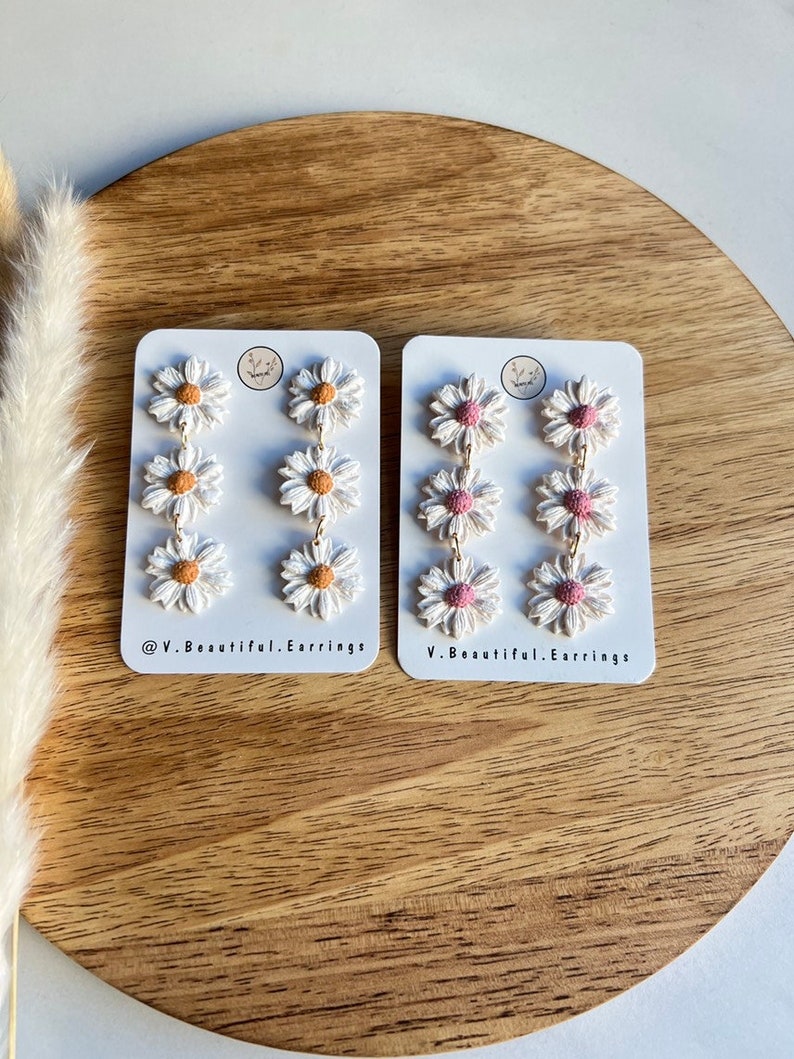 May include: Two pairs of daisy earrings on white cards. The earrings are made of white polymer clay with a pink or orange center. The cards have the text "@V.Beautiful.Earrings" and "V.Beautiful.Earrings".