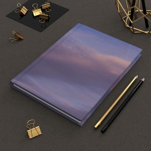 May include: A purple notebook with a cloudy sky design on the cover. The notebook is lying on a grey surface with two pencils and gold paper clips.