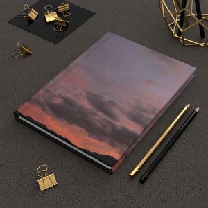 May include: A closed hardback journal with a sunset sky image on the cover. The journal is lying on a grey textured surface with two pencils and gold paper clips.