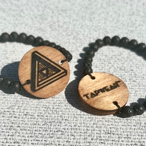 Può includere: Due braccialetti con perline di lava nera e ciondoli in legno. Un ciondolo ha un design geometrico e l'altro dice "TAPWEAR".