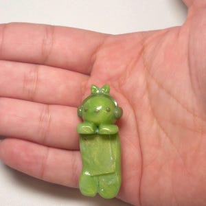 Pu&ograve; includere: Piccola statuina verde di un personaggio dei cartoni animati con cuffie e fiocco. La statuina ha un corpo rettangolare e tiene le mani unite. La statuina &egrave; realizzata in un materiale traslucido e lucido.