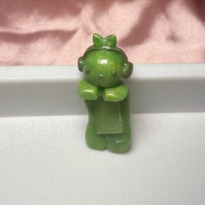 Pu&ograve; includere: Piccola statuina verde di un personaggio con un fiocco sulla testa e delle cuffie. La statuina &egrave; in posizione eretta con le mani giunte davanti. La statuina &egrave; su una superficie bianca.