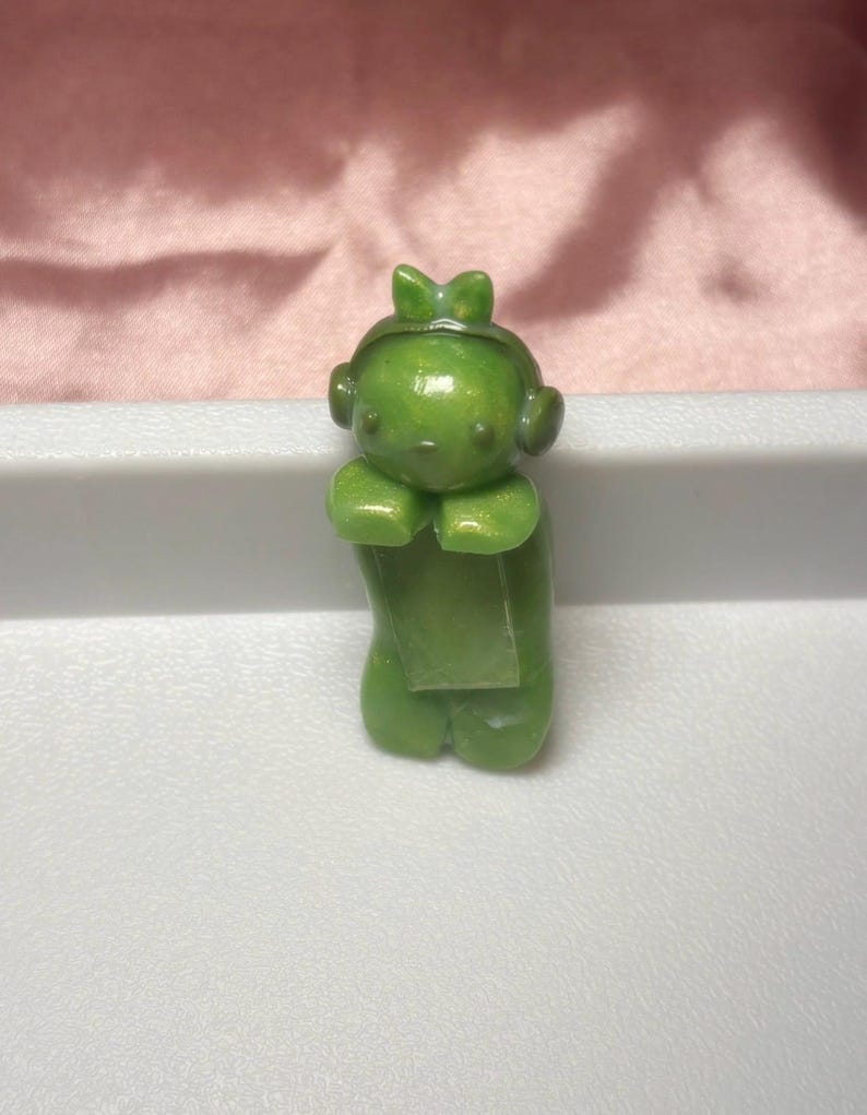 Pu&ograve; includere: Piccola statuina verde di un personaggio con un fiocco sulla testa e delle cuffie. La statuina &egrave; in posizione eretta con le mani giunte davanti. La statuina &egrave; su una superficie bianca.