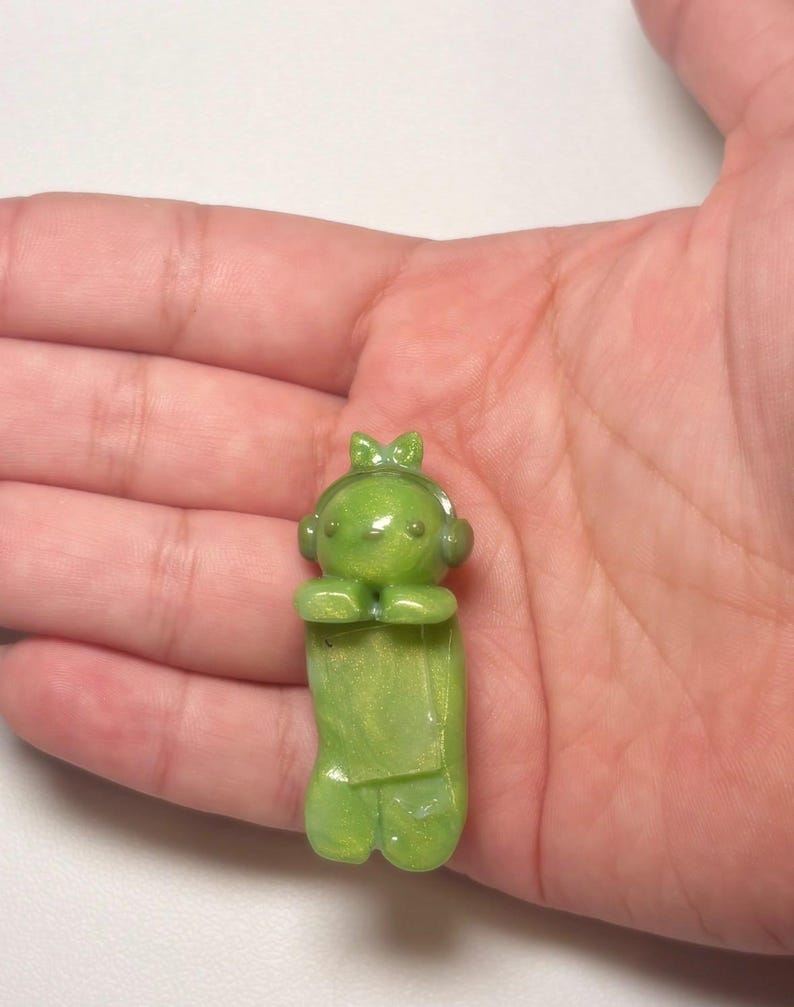 Pu&ograve; includere: Piccola statuina verde di un personaggio dei cartoni animati con cuffie e fiocco. La statuina ha un corpo rettangolare e tiene le mani unite. La statuina &egrave; realizzata in un materiale traslucido e lucido.
