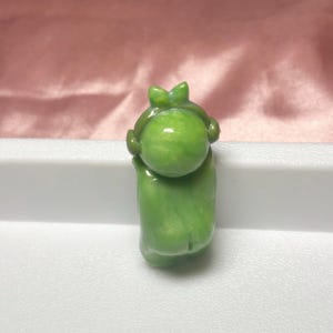 Pu&ograve; includere: Piccola statuetta verde raffigurante una persona con la testa rotonda e un fiocco. La statuetta tiene le braccia alzate e ha una finitura liscia e lucida. Lo sfondo &egrave; un tessuto rosa tenue.
