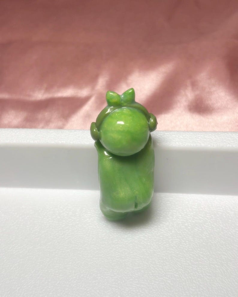 Pu&ograve; includere: Piccola statuetta verde raffigurante una persona con la testa rotonda e un fiocco. La statuetta tiene le braccia alzate e ha una finitura liscia e lucida. Lo sfondo &egrave; un tessuto rosa tenue.