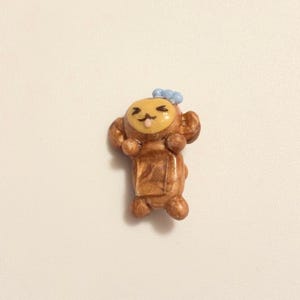 May include: A small, handmade figurine of a monkey with a light brown body, a yellow face, and a blue bow. The monkey has its arms raised and a small tongue sticking out. The figurine is on a white background.