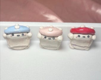 May include: Three small, whimsical figurines with mushroom-shaped hats in blue, pink, and red. The figurines have white bodies and small, round faces with black eyes and a small mouth. They are arranged on a white surface.
