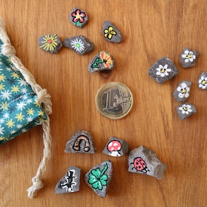 Könnte beinhalten: Bemalte Steine mit floralen und skurrilen Designs, ein türkisfarbener Stoffbeutel mit Sternmuster und eine Ein-Euro-Münze. Die Steine sind mit Blumen, einem Pilz, einem Marienkäfer und anderen Symbolen bemalt.