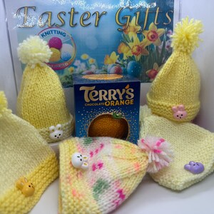 May include: A collection of handmade yellow knitted baby hats with pom-poms and bunny embellishments, alongside a box of Terry's Chocolate Orange. The hats are arranged with a matching scarf, and the background features Easter-themed decorations.