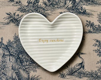 Może przedstawiać: Biała ceramiczna miseczka w kształcie serca z żebrowaną fakturą i napisem "Enjoy sunshine" wykonanym złotymi literami na środku. Naczynie ma gładkie, błyszczące wykończenie oraz wyraźny kształt serca ze spiczastym dołem i zaokrągloną górą.