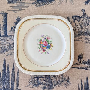 Peut inclure: Une assiette carrée en céramique couleur crème aux coins arrondis, ornée d'un bouquet floral central aux tons rose, bleu, jaune et vert. Le bord est décoré d'un liseré doré et d'une texture en relief rappelant une corde sur le pourtour.