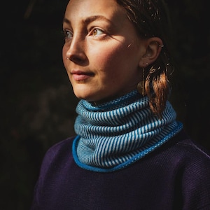 May include: A person wearing a blue and white striped neck warmer with a blue trim. The neck warmer is made of a soft, knitted fabric. The person is wearing a dark purple sweater.