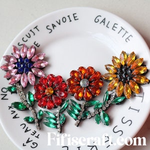 May include: Five colorful rhinestone flower embellishments with green rhinestone leaves. The flowers are pink, red, orange, and yellow. The flowers are arranged on a white plate with the words "Galeta Savoie" written on the plate.