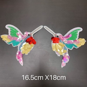 Puede incluir: Dos apliques de colibrí iridiscentes con lentejuelas y cuentas. Los colibríes miden 16,5 cm por 18 cm.