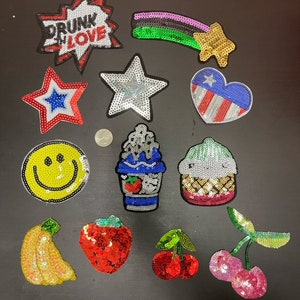 Op de afbeelding: Assortiment van kleurrijke strijkbare patches met verschillende ontwerpen, waaronder een ster, een hart, een smiley, een banaan, een aardbei, een kers, een ijshoorn, een cupcake en een vallende ster. De patches zijn gemaakt van pailletten en hebben een glanzende afwerking.
