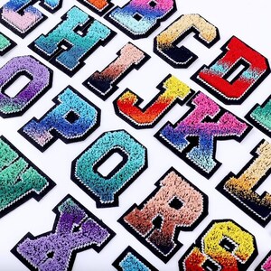 May include: A set of 26 colorful, fuzzy, iron-on alphabet patches. Each letter is outlined in black and has a gradient of colors. The letters are arranged in alphabetical order.